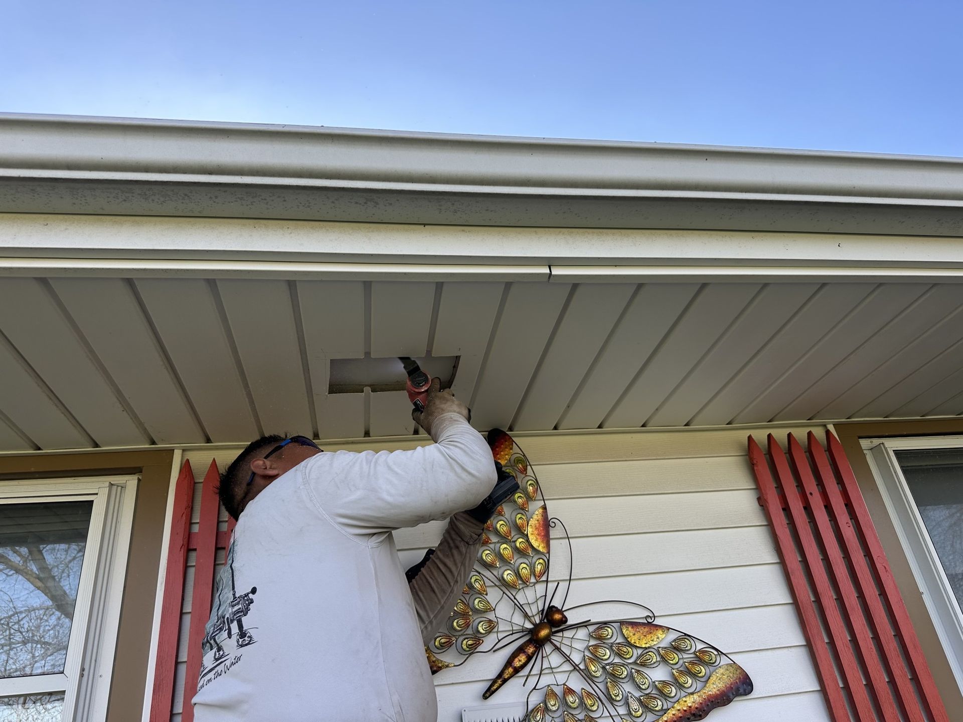 A person in a light-colored shirt works on the exterior lighting fixture under the eaves of a house with siding.