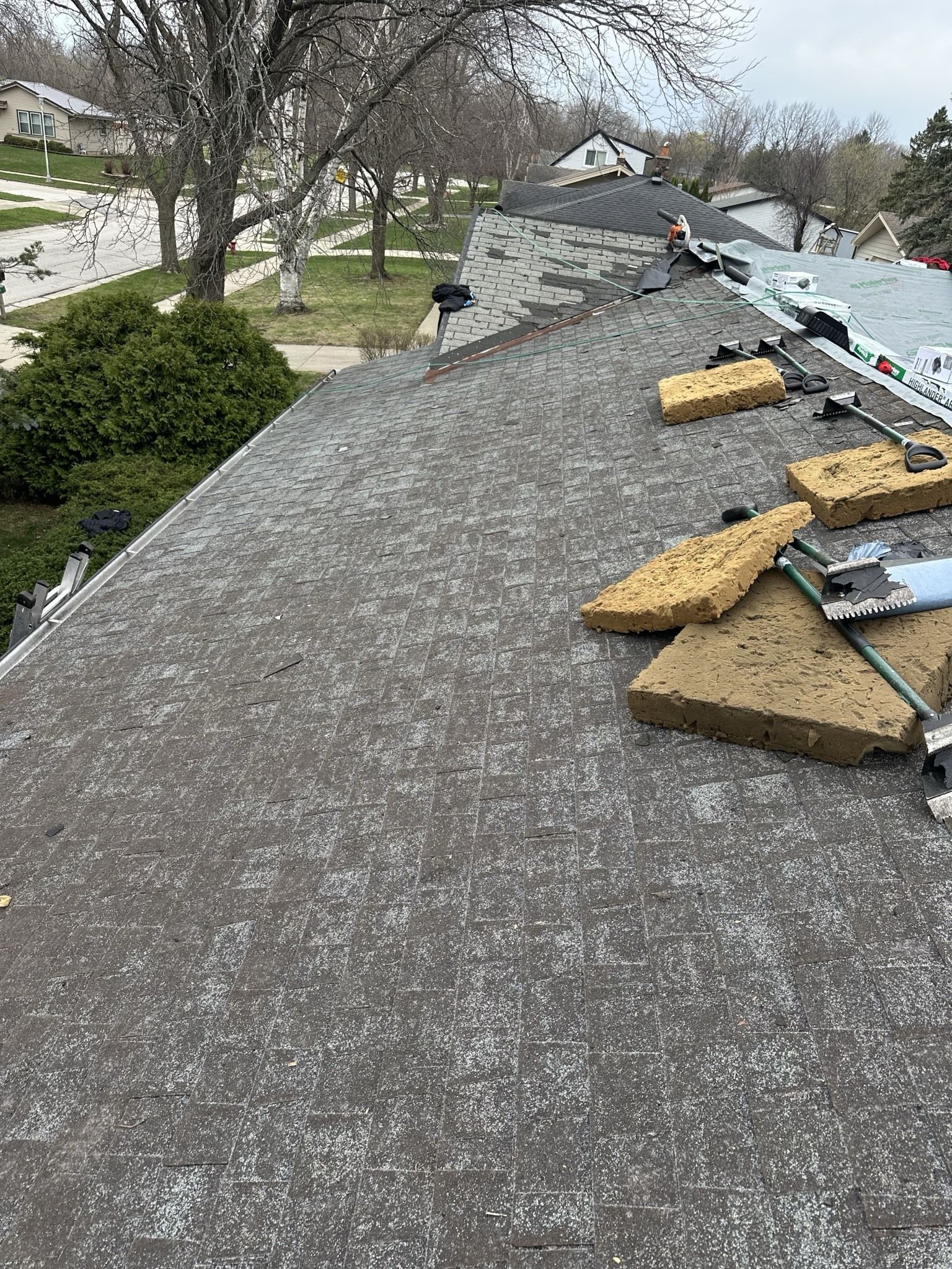 A view of a roof under construction, featuring exposed roofing material, stacked insulation blocks, and a suburban street.