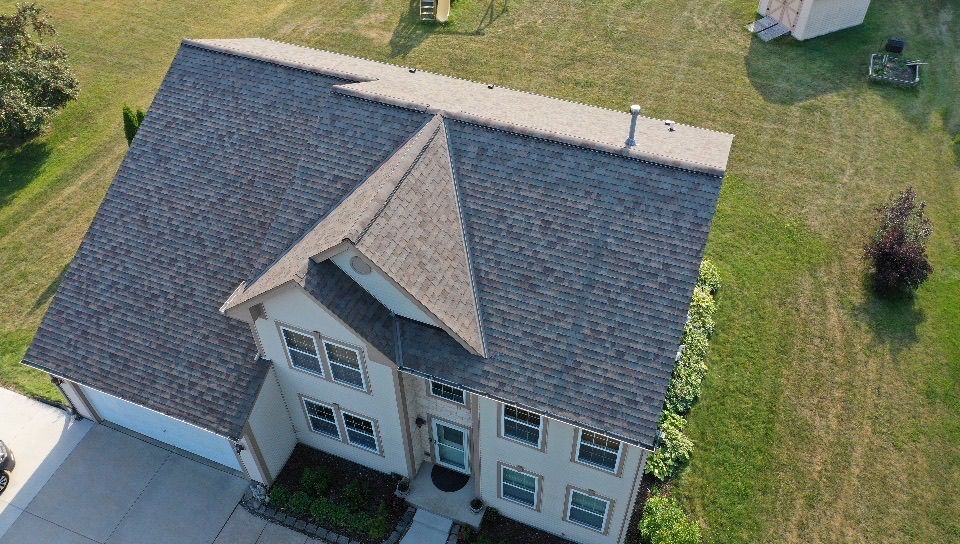 An aerial view of a two-story house with a grey shingled roof, beige siding, and an attached garage on a grassy lot.