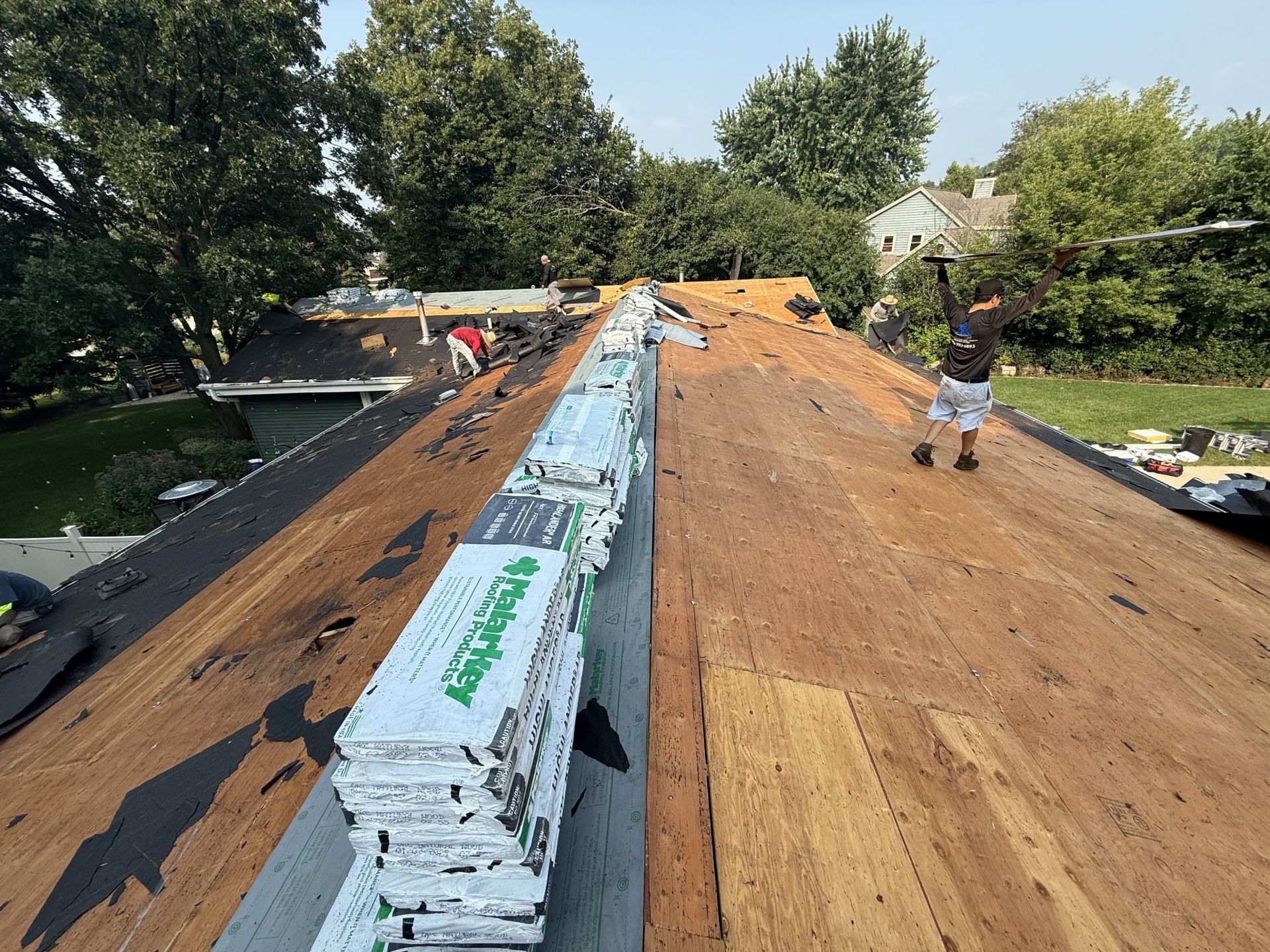 Construction workers install roofing materials on a partially stripped residential house roof on a sunny day.