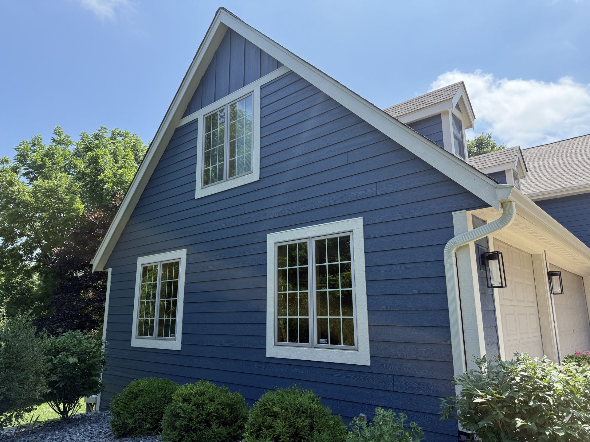 A two-story blue house with white trim and a gabled roof set against a blue sky with lush green trees and bushes nearby.