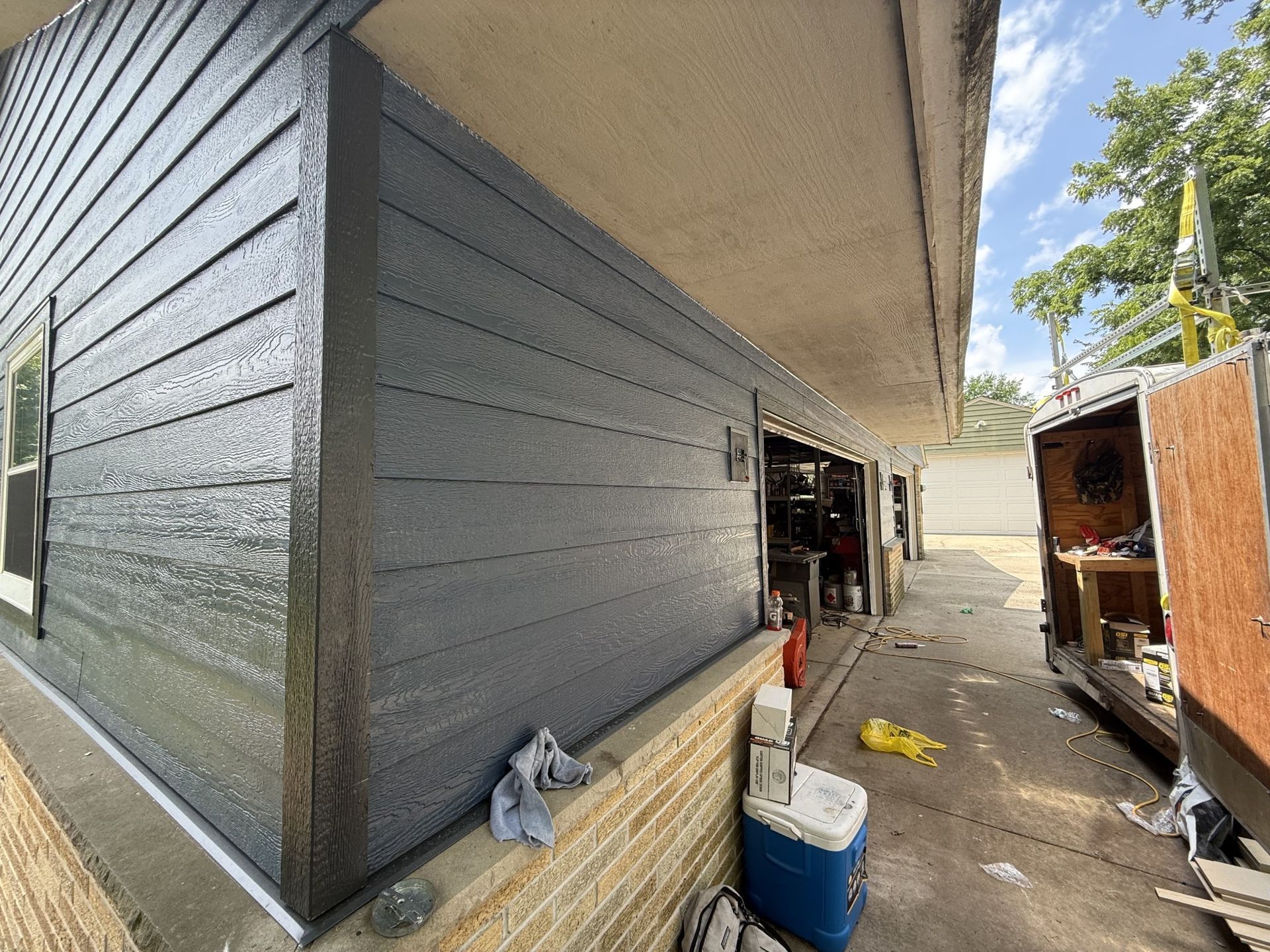 A house exterior showing dark gray horizontal siding above a brick base, next to a driveway with a trailer.