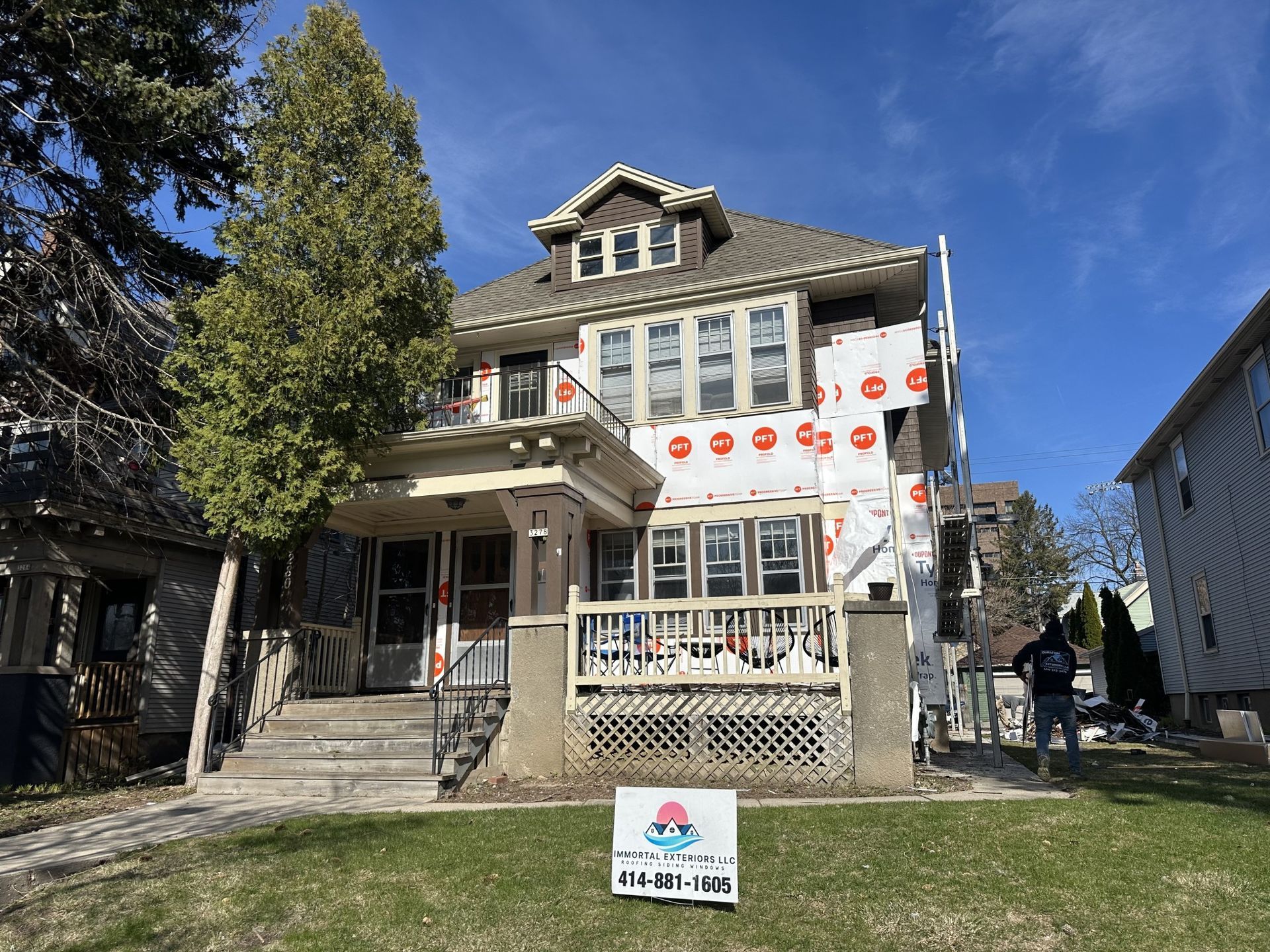A large house under renovation with protective wrap on the siding, a wooden porch, and a construction sign in the yard.