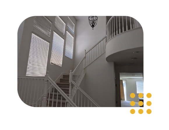 A white interior staircase with a curved landing, wood railings, and tall windows with blinds in a high-ceilinged foyer.