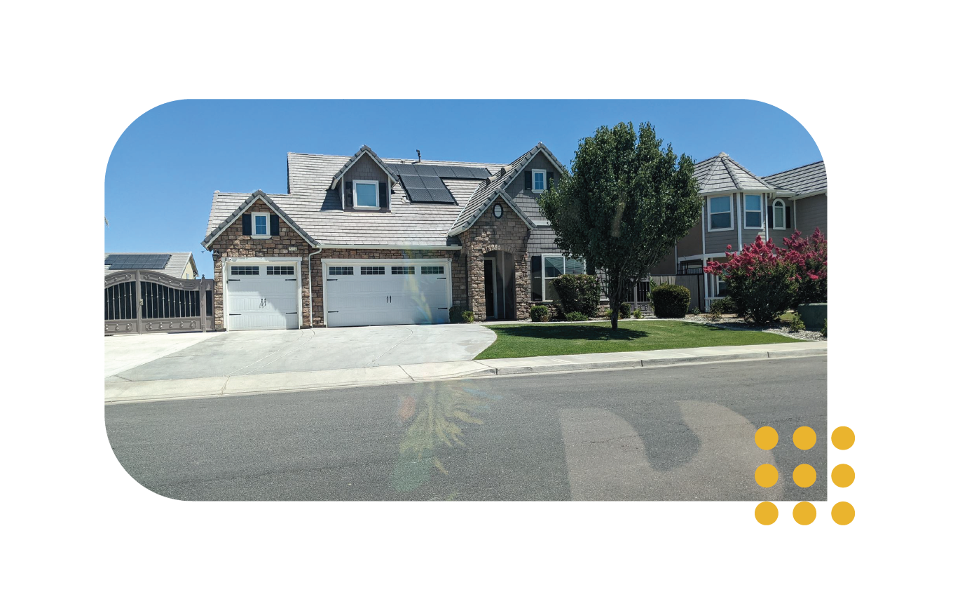 A two-story suburban house with a stone facade, a two-car garage, solar panels on the roof, and a front yard with a tree.