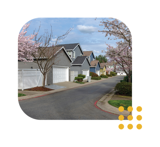 Quiet suburban street with gray houses, white garages, and blooming pink trees under a blue sky
