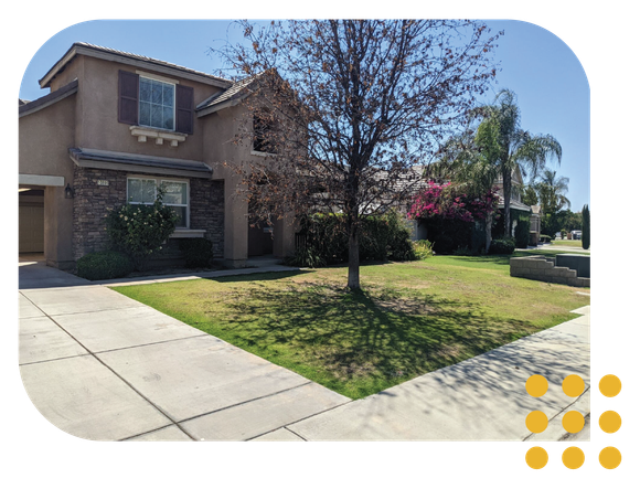 A two-story tan suburban house with a stone facade, a concrete driveway, and a front yard with a tree and bushes.