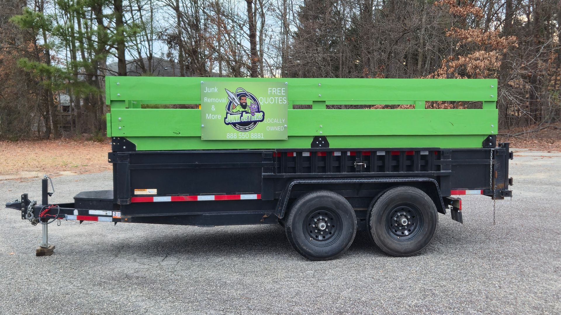 A green and black dump trailer with a company logo on the side parked on a gravel lot in front of trees.