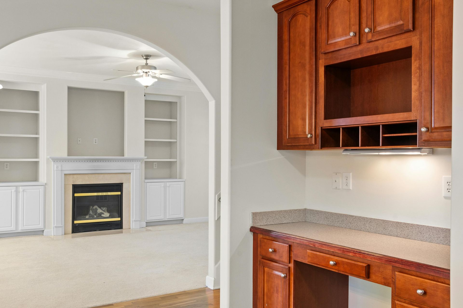 Living room and built-in desk with wood cabinets. Fireplace, archway, and neutral colors.