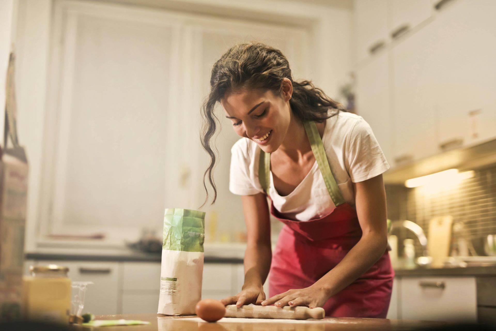 Woman in apron rolling dough in kitchen, smiling. Flour and egg on counter.