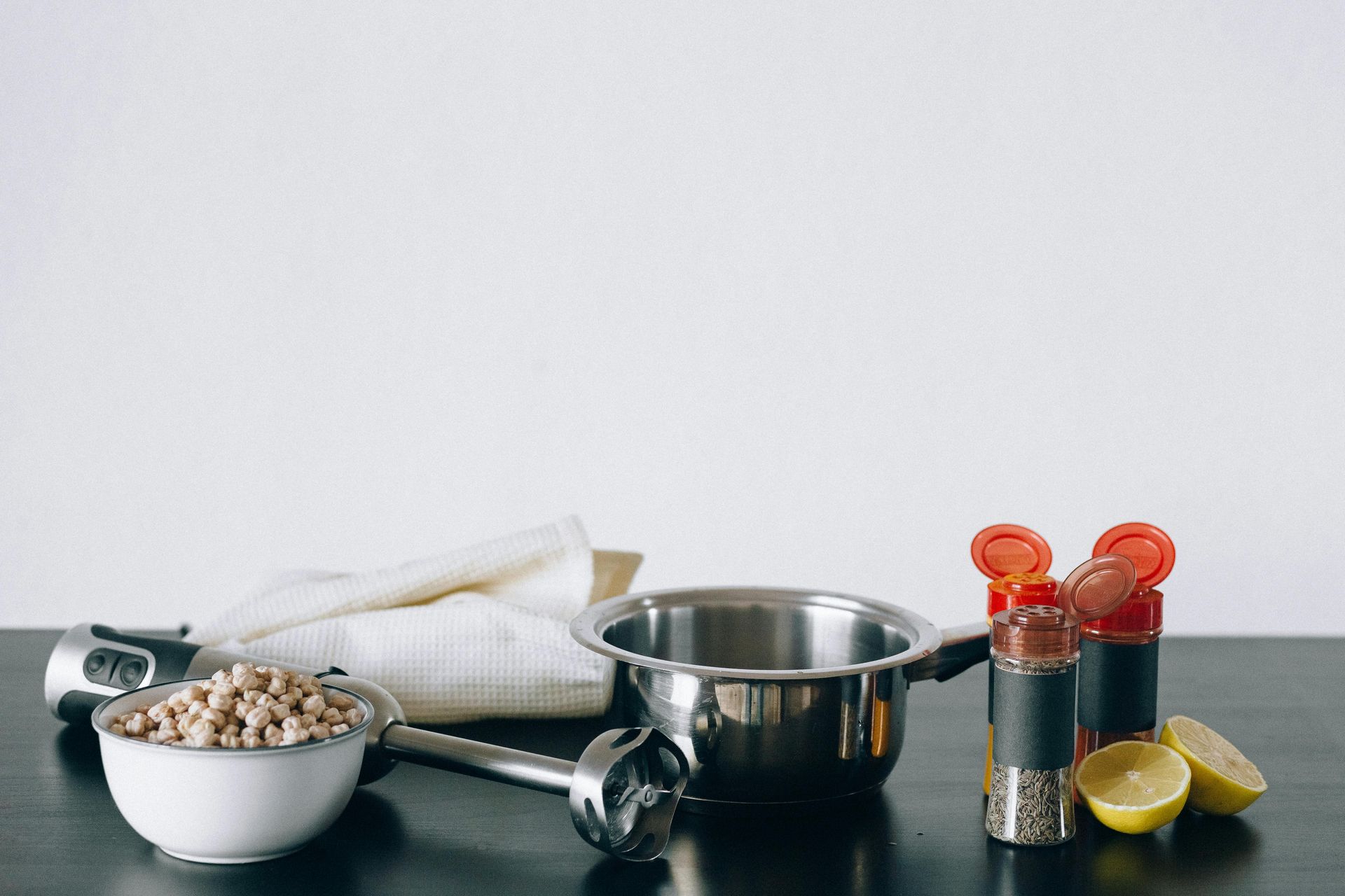 Bowl of chickpeas, immersion blender, pot, lemons, and spices on a black surface.