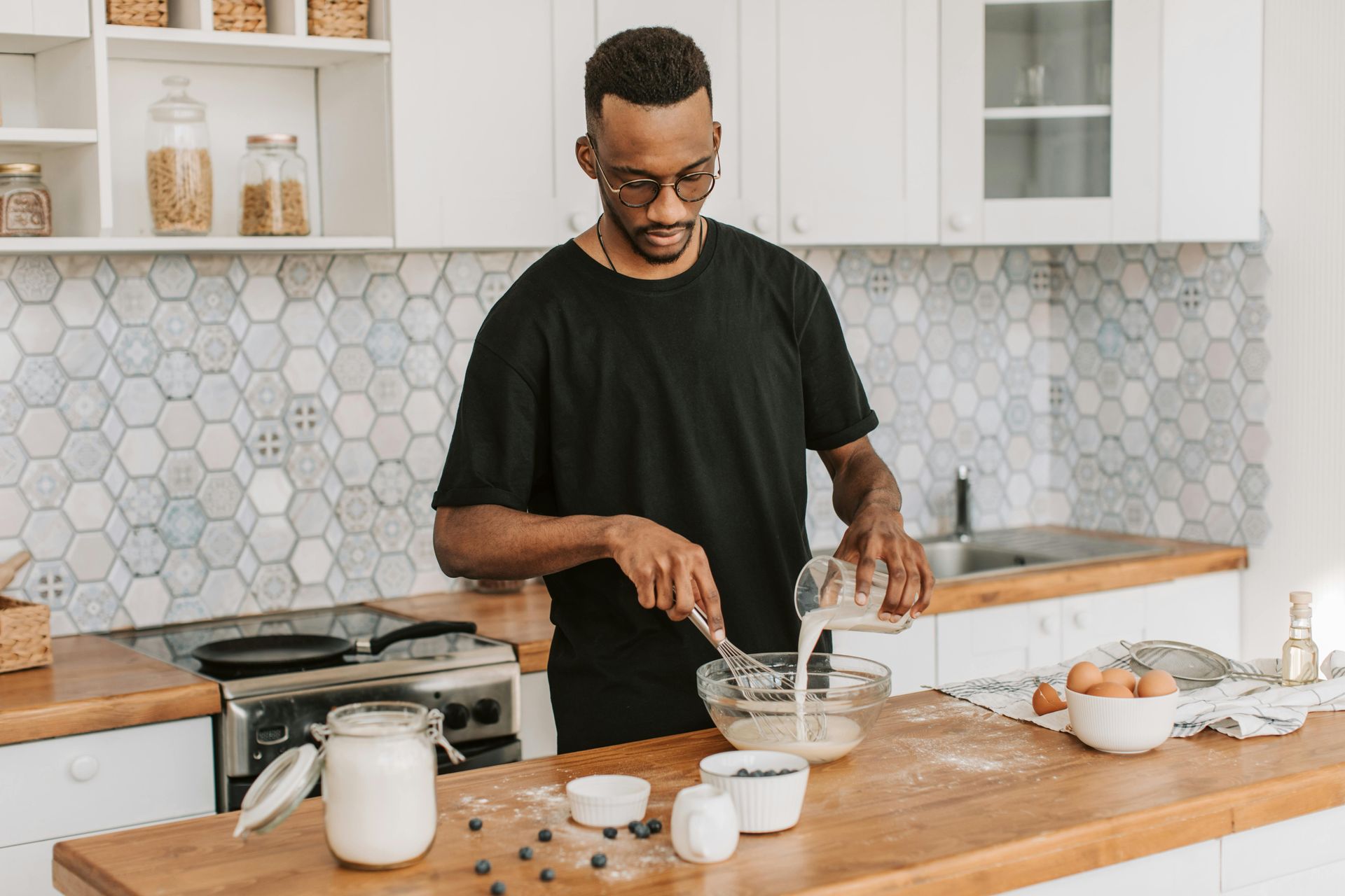 Man in black shirt cooks in bright kitchen, pouring milk into a bowl while whisking ingredients.