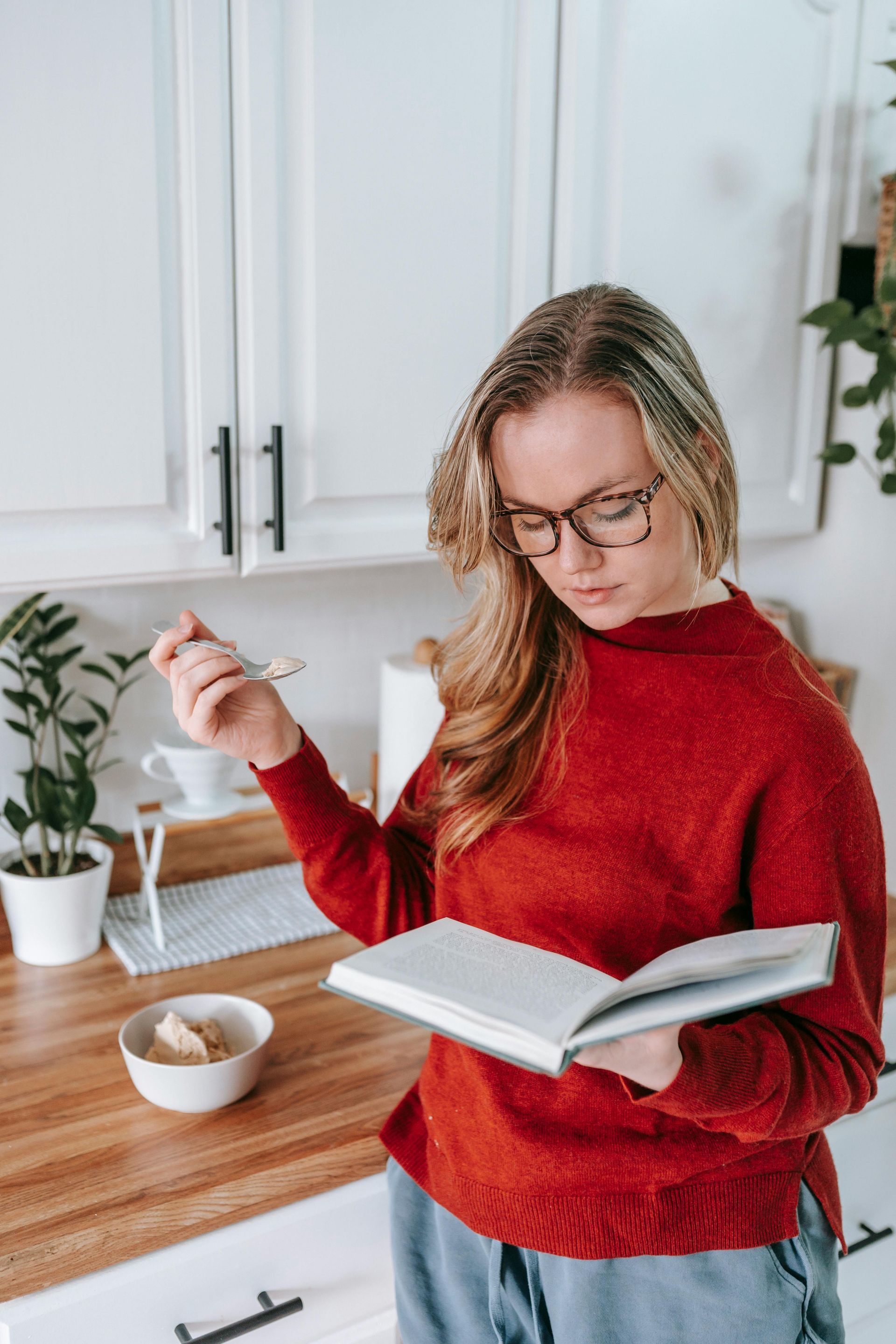 Woman in red sweater and glasses in a kitchen reading a book, holding a fork near a bowl of food.