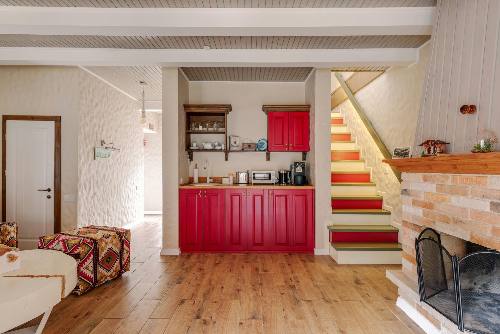 Cozy living space with red kitchen cabinets, stairs, and fireplace. Light wood floors, white walls.