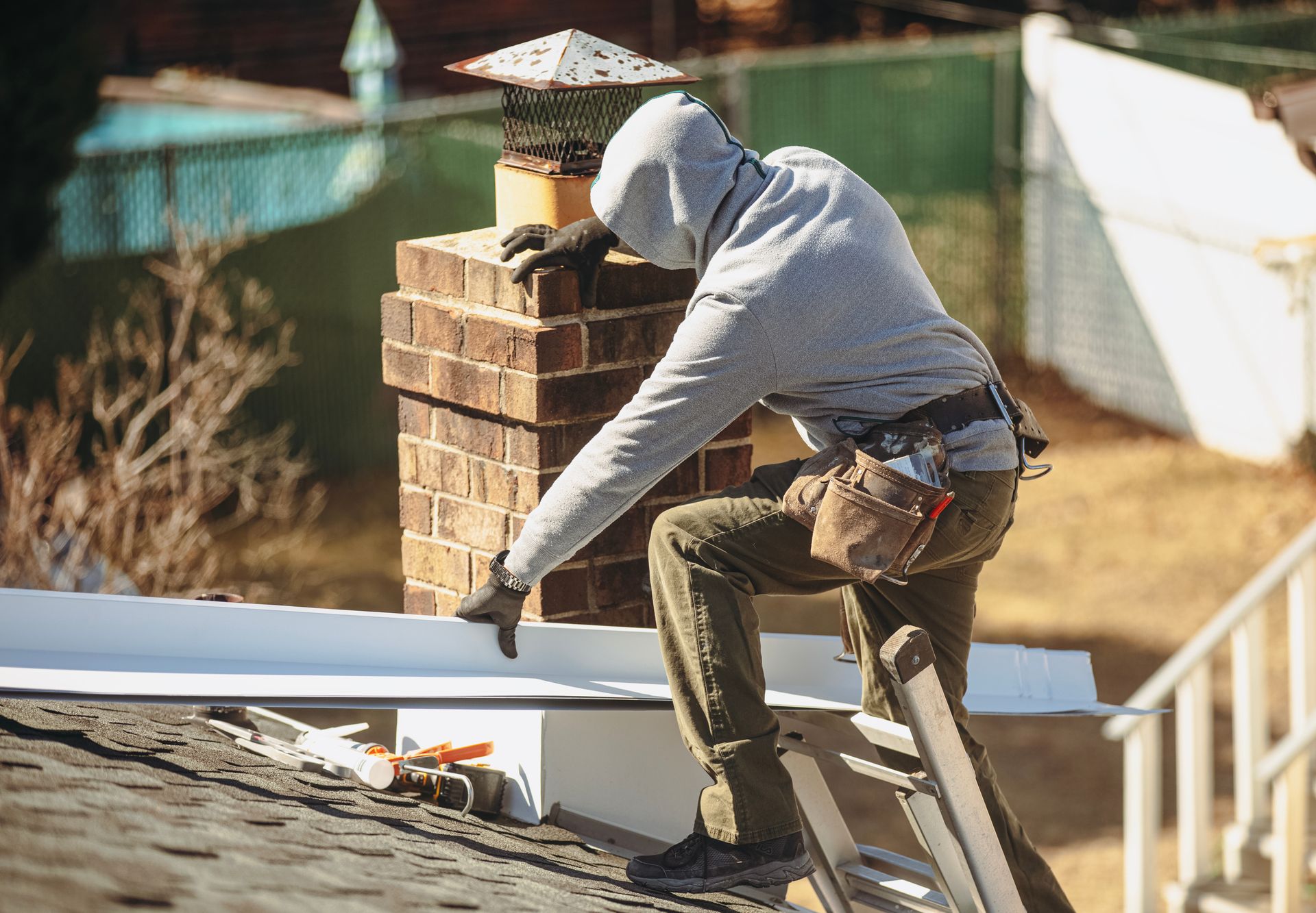 Person in hooded sweatshirt on a roof, installing flashing around a brick chimney.