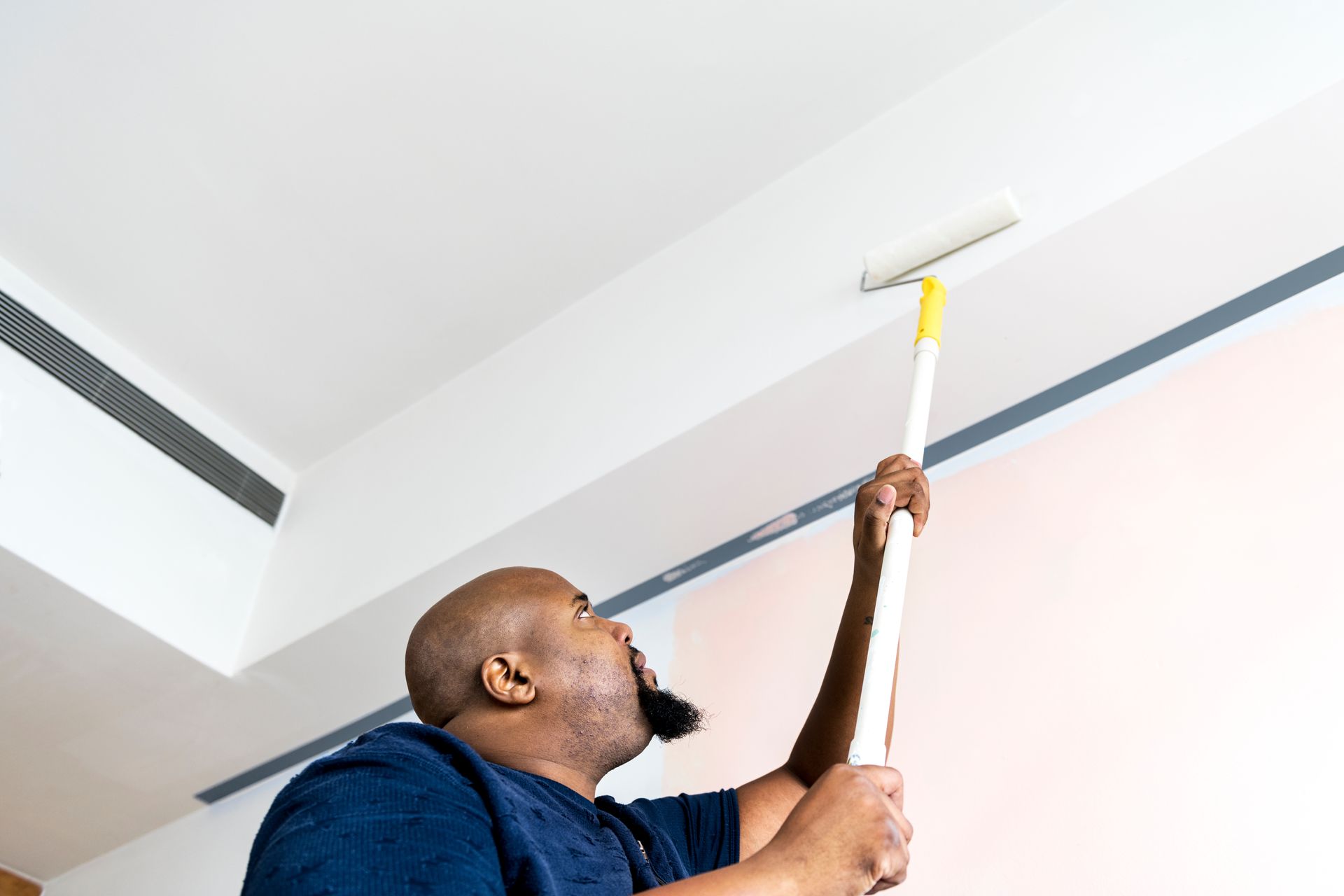 A Black man paints a ceiling with a roller. He's looking up with mouth slightly open.