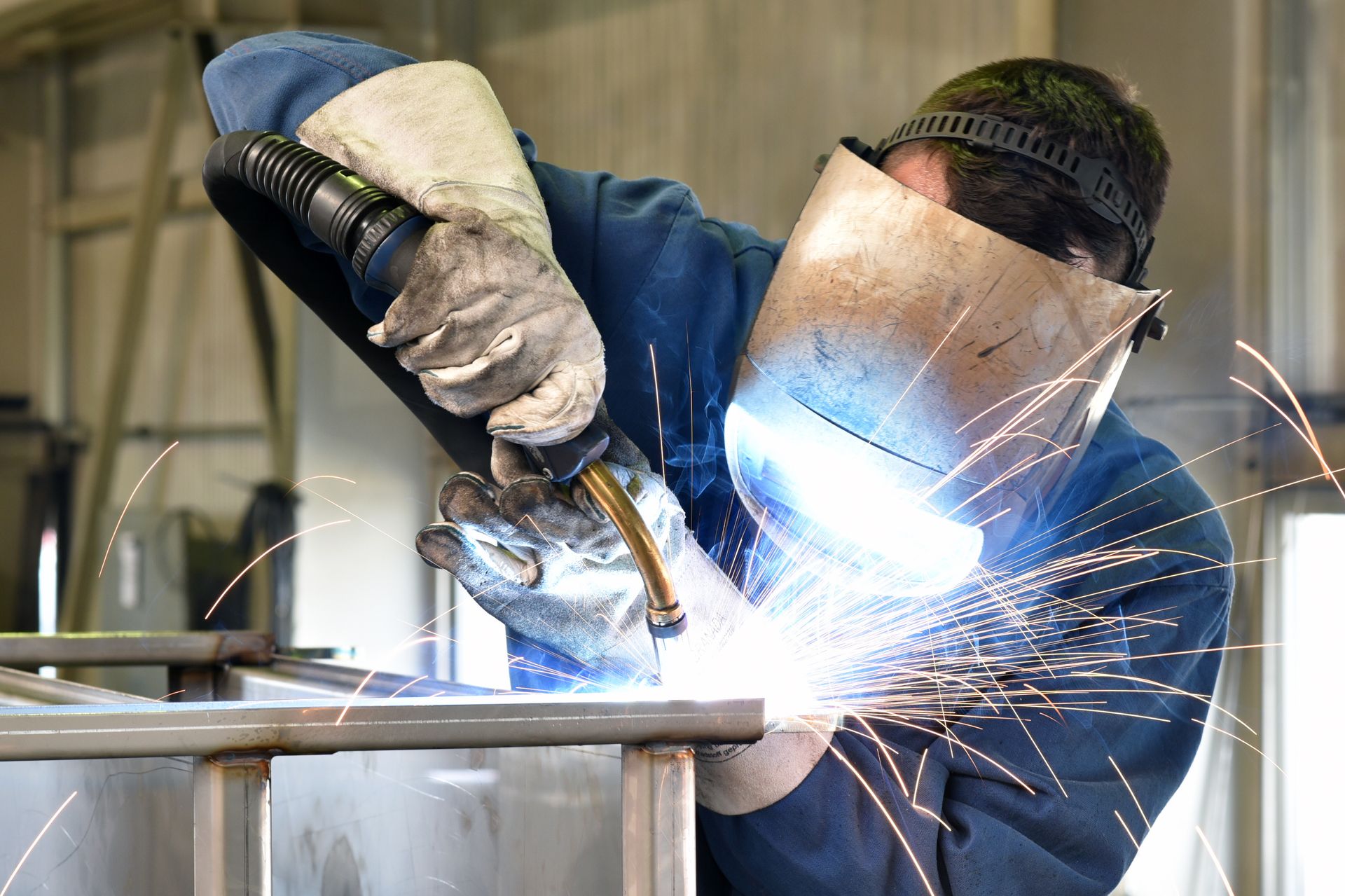 Welder wearing safety gear, welding metal with bright sparks in a workshop.