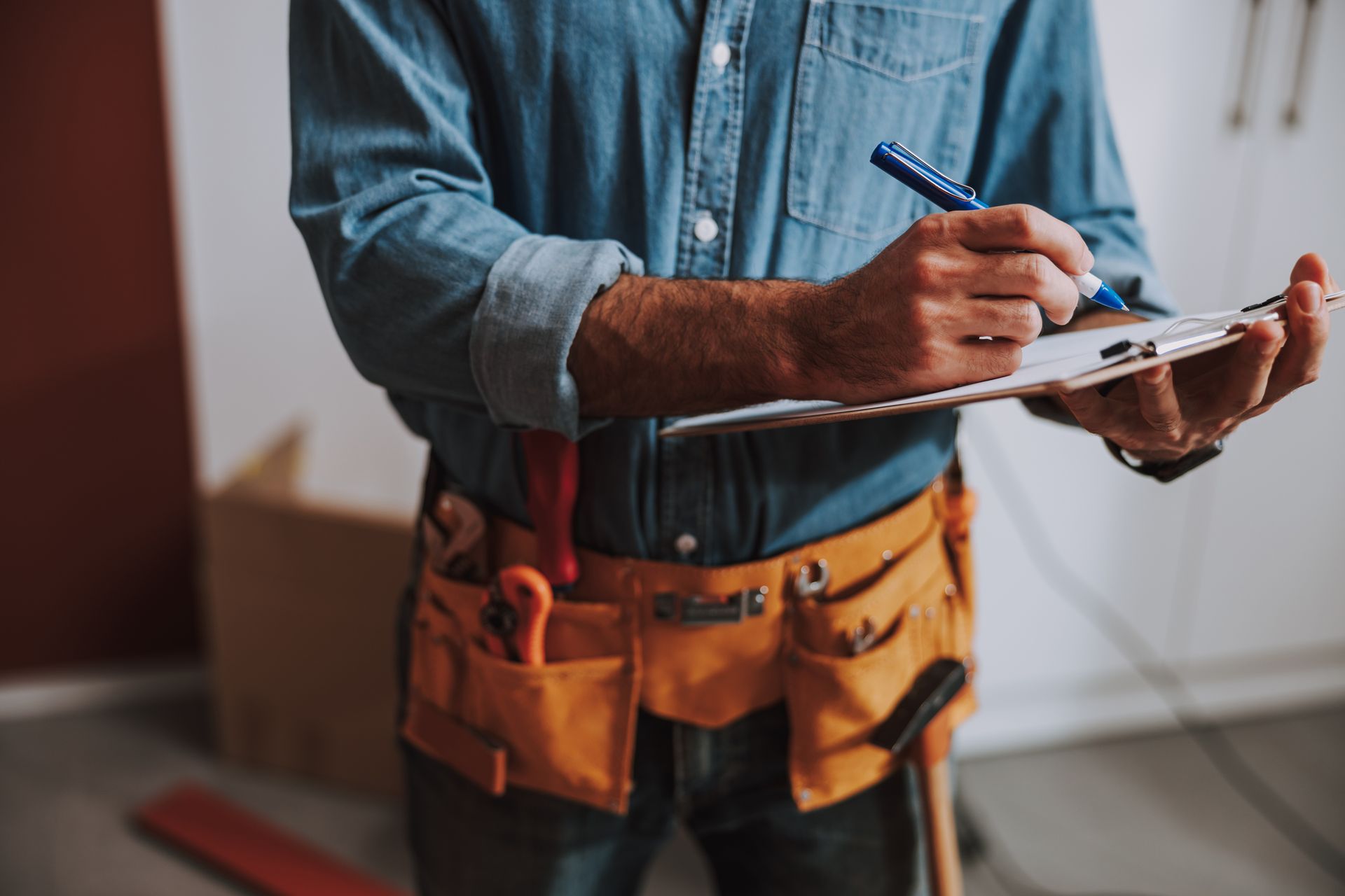 A person in a blue shirt with a tool belt, writing on a clipboard indoors.
