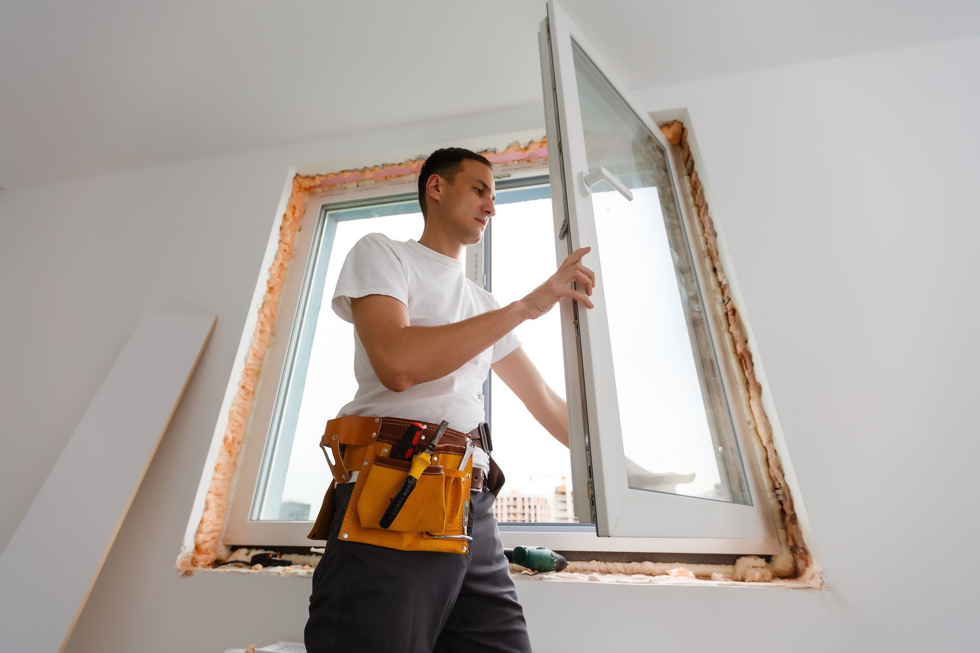 Man installing a window in a white room, wearing a tool belt.