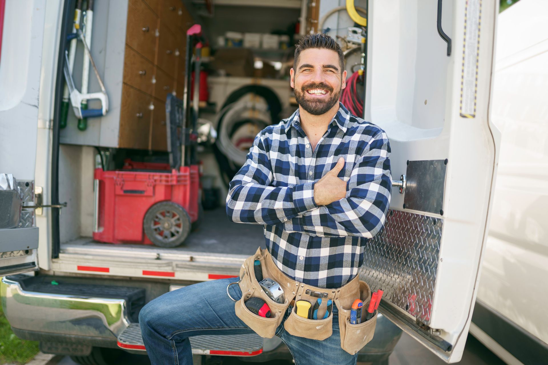 Smiling handyman in plaid shirt with tools, leaning against open van.