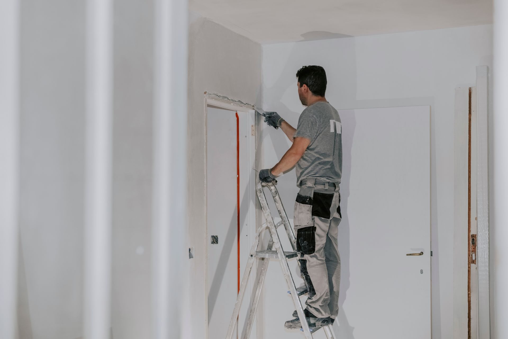 Painter on a ladder, applying paint to a doorway. White walls, neutral tones.