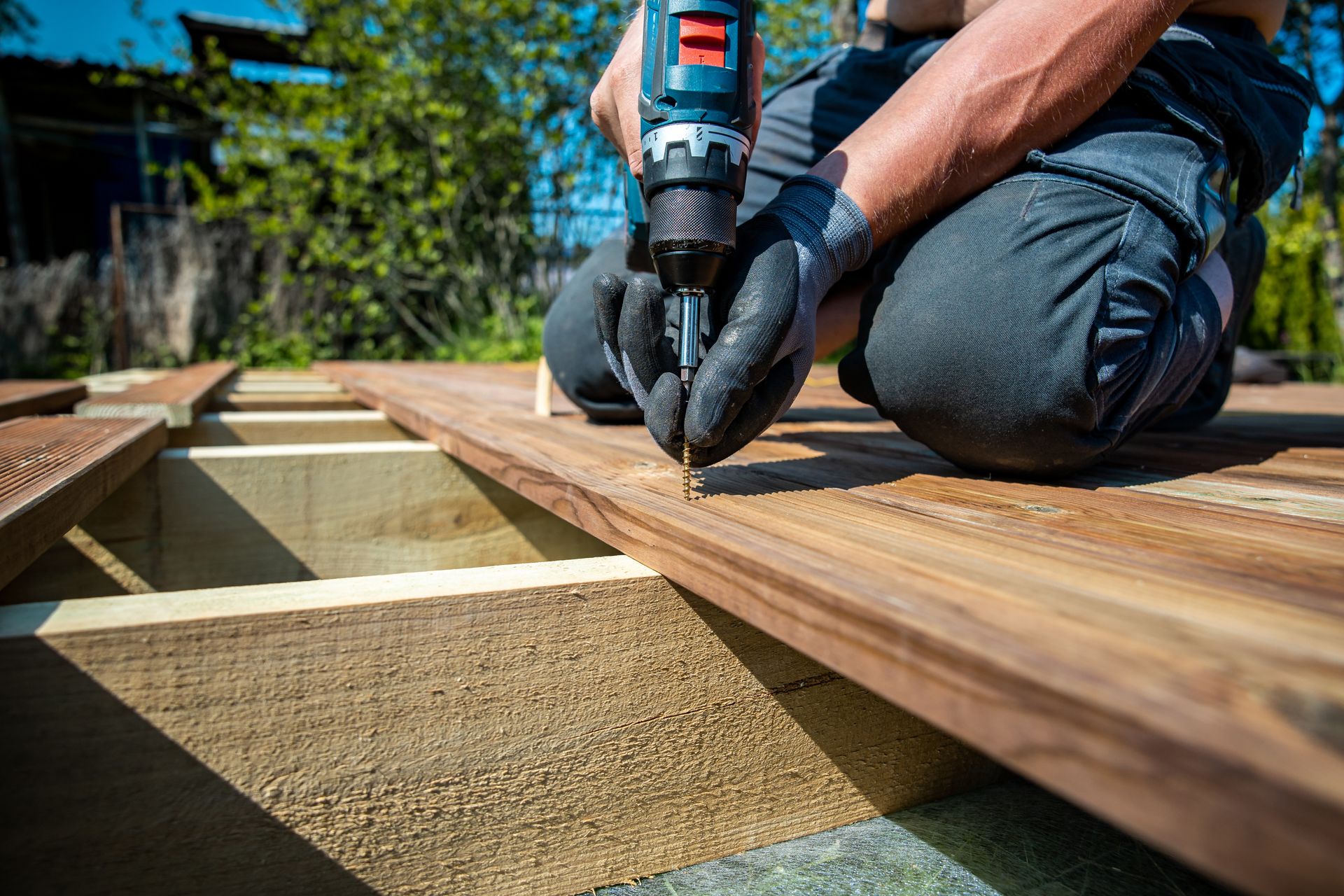 Person kneeling, using a power drill to screw a wooden deck board in place outdoors.