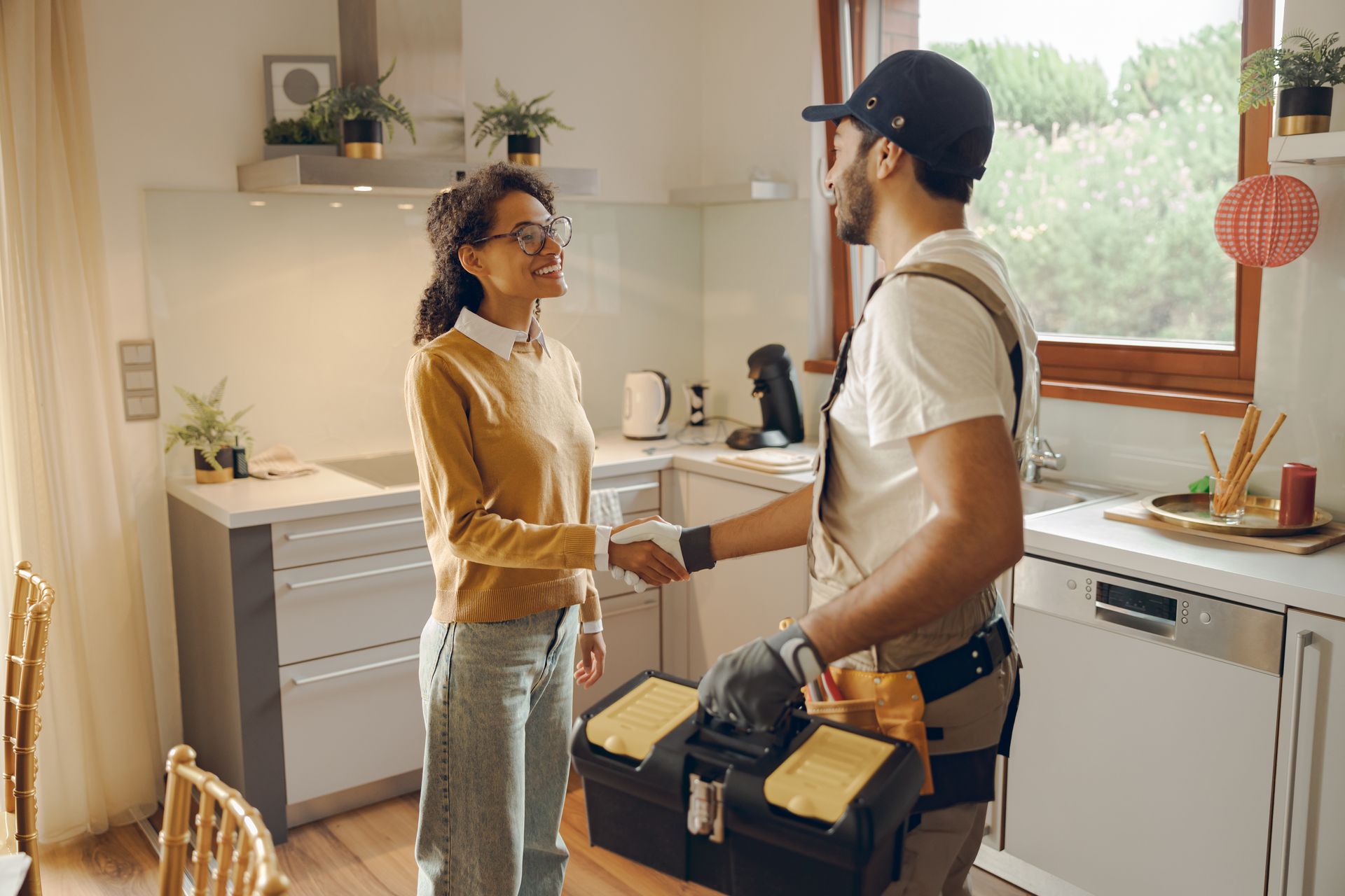 Woman shakes hands with a tradesperson in a kitchen; smiling, toolbox present.