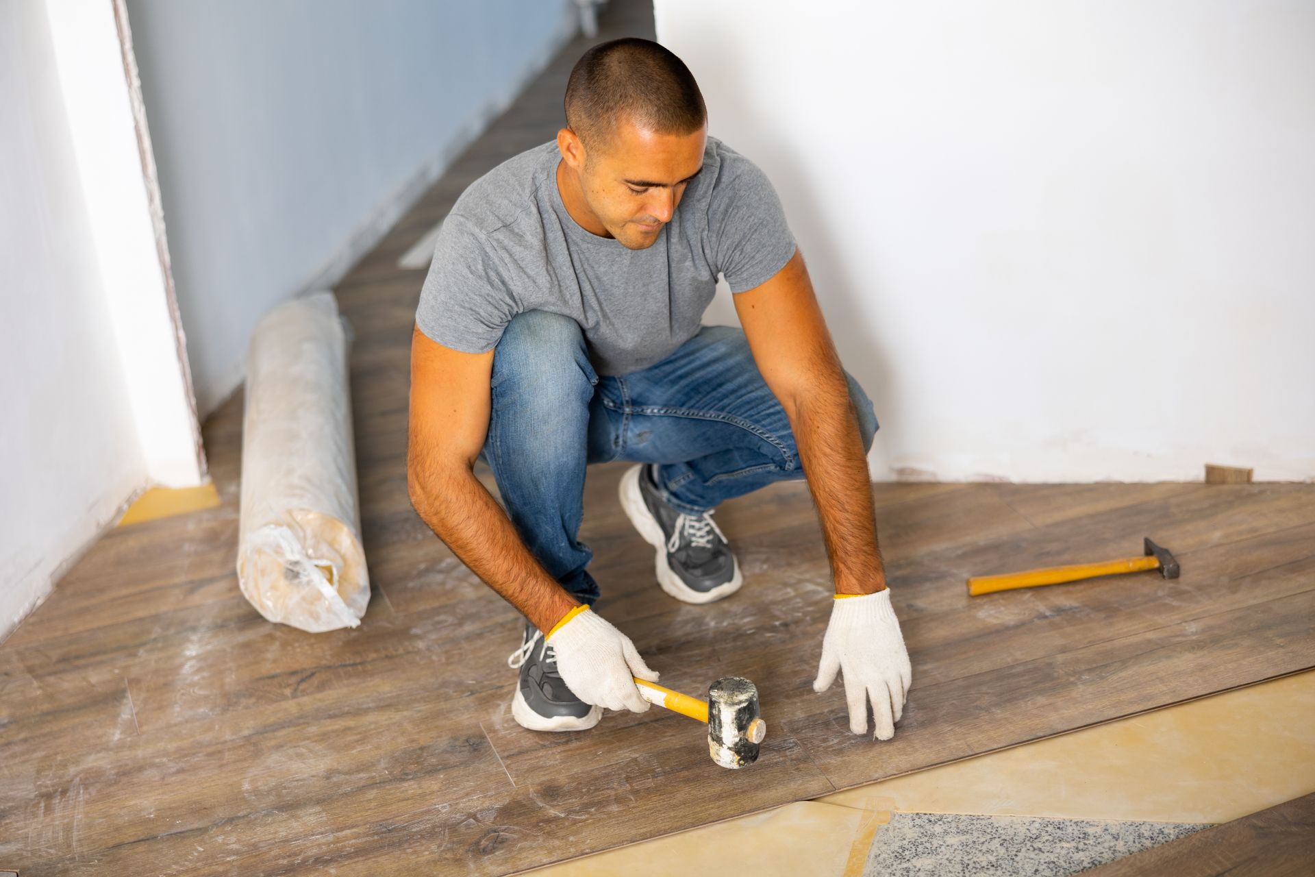 Man installing floorboards, wearing gloves, using a hammer. Inside a room.