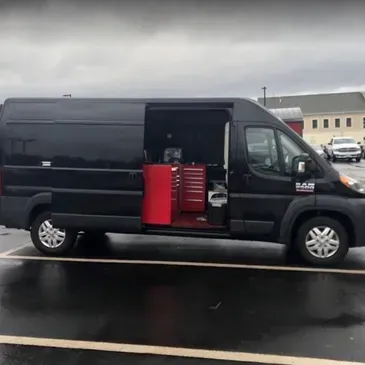 Black cargo van with side door open, showing red tool cabinets inside, parked on a wet road.