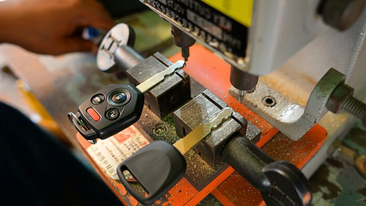 Car keys clamped in a metal vise under a machine tool, with a key fob and metal key visible
