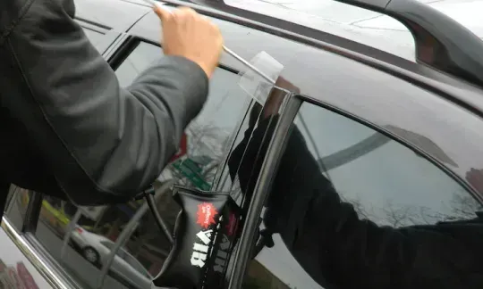 Person using a tool to unlock a black car door through the window seal