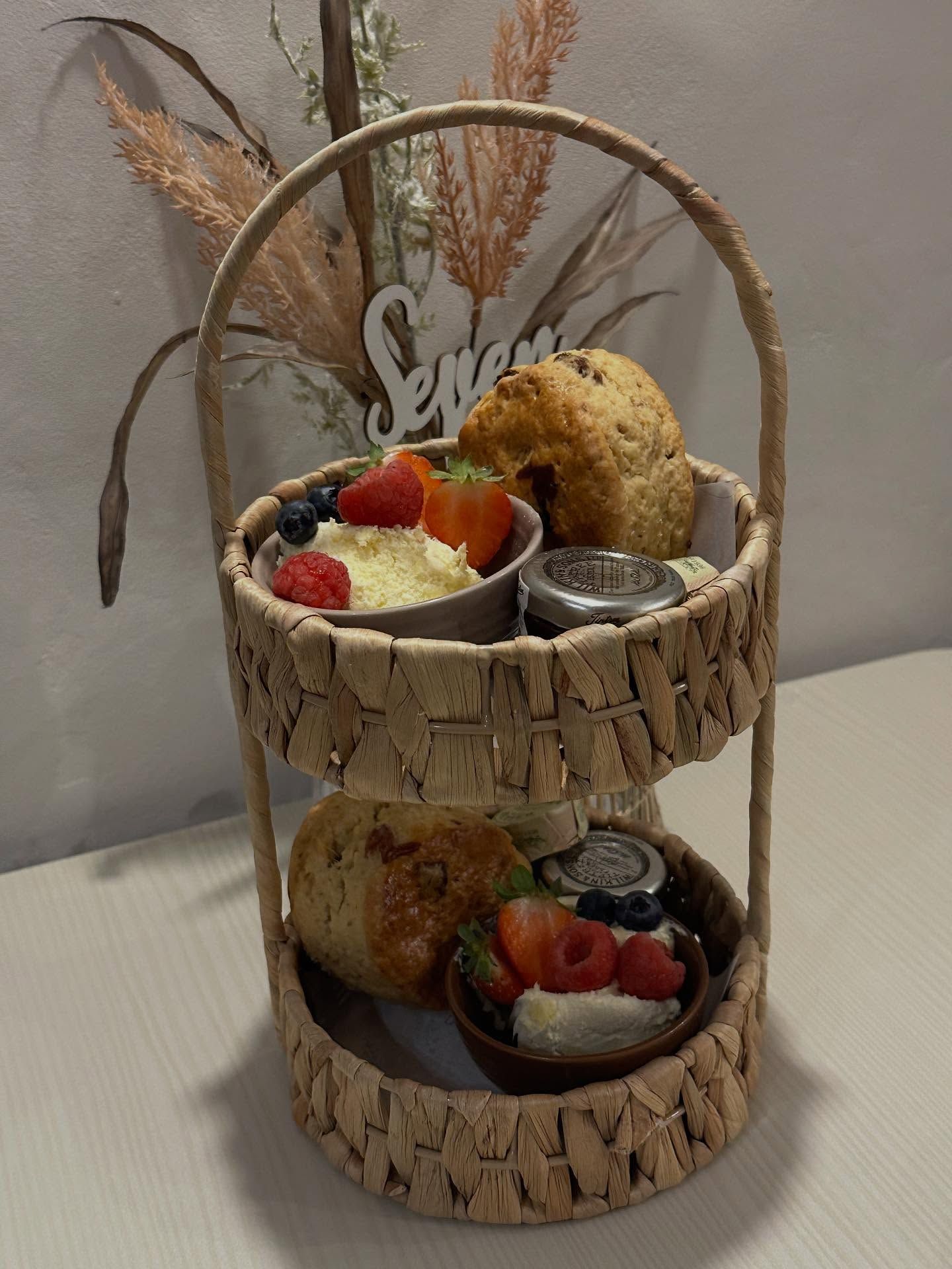 Two-tiered woven basket filled with treats like scones, fruit, and cream, on a table.