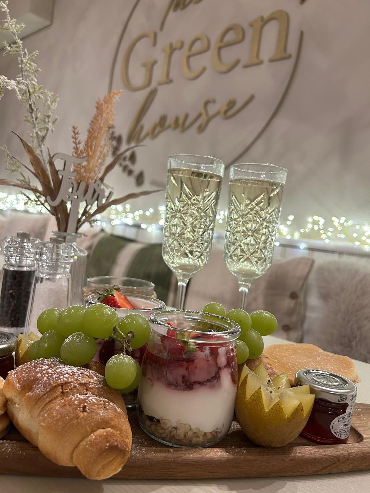 Charcuterie board with champagne, grapes, pastries, and a dessert jar; in front of a sign that reads 