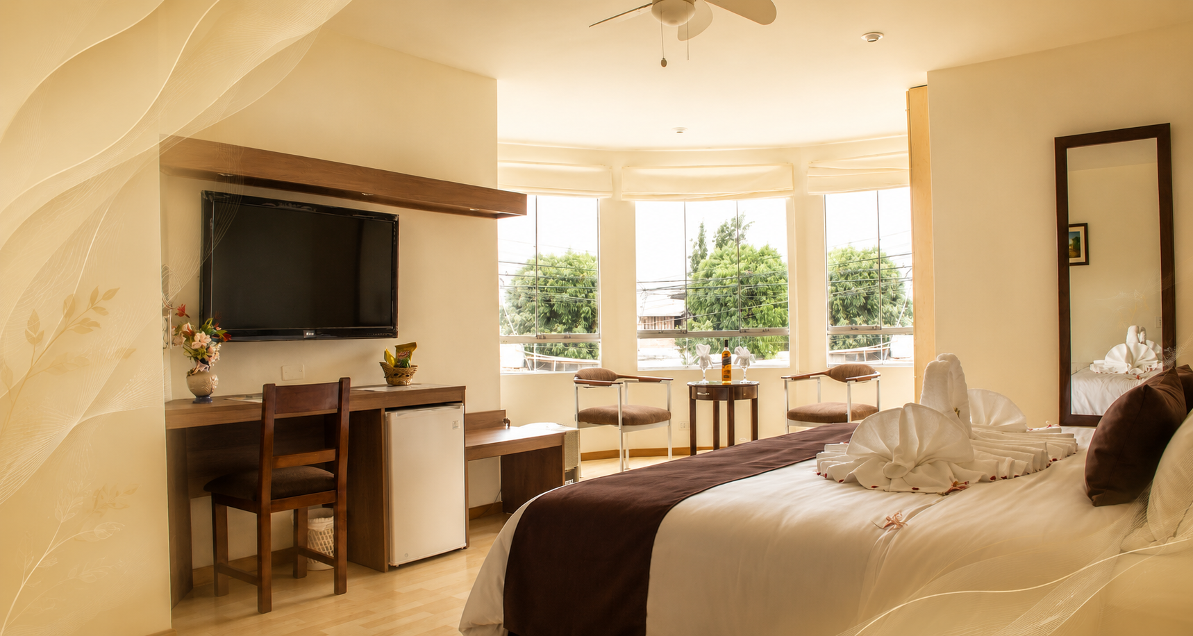 Habitación de hotel luminosa con cama, escritorio, televisor y grandes ventanales con vistas a zonas verdes.
