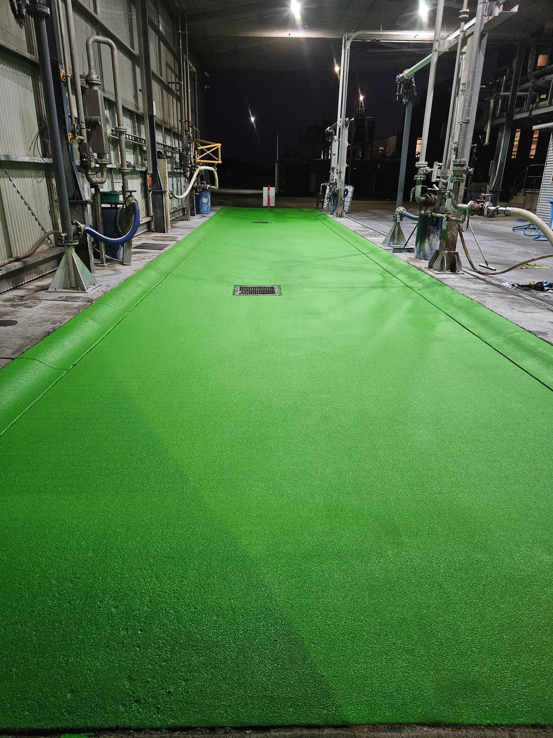 Green synthetic turf covering an industrial floor. Long, narrow area with drains, metal structures, and dark background — Riverina Epoxy Flooring In Glenfield Park, NSW