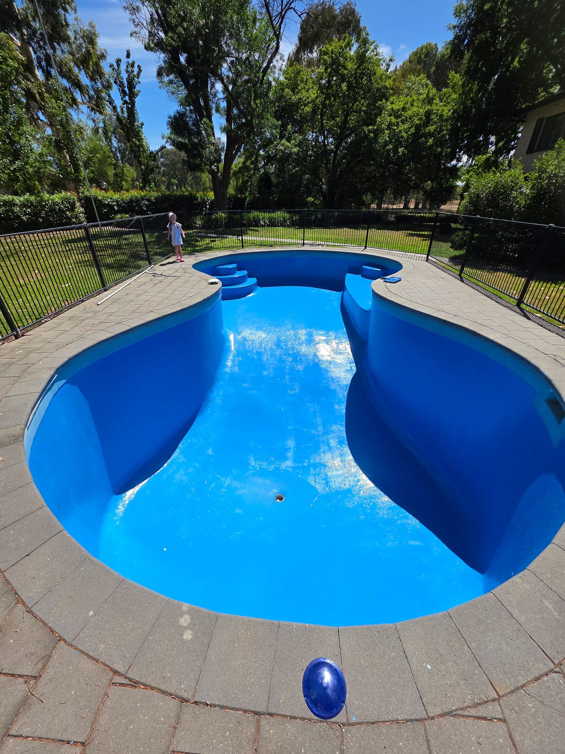 Blue-painted swimming pool surrounded by gray stone paving. A child stands in the background. Green trees — Riverina Epoxy Flooring In Glenfield Park, NSW