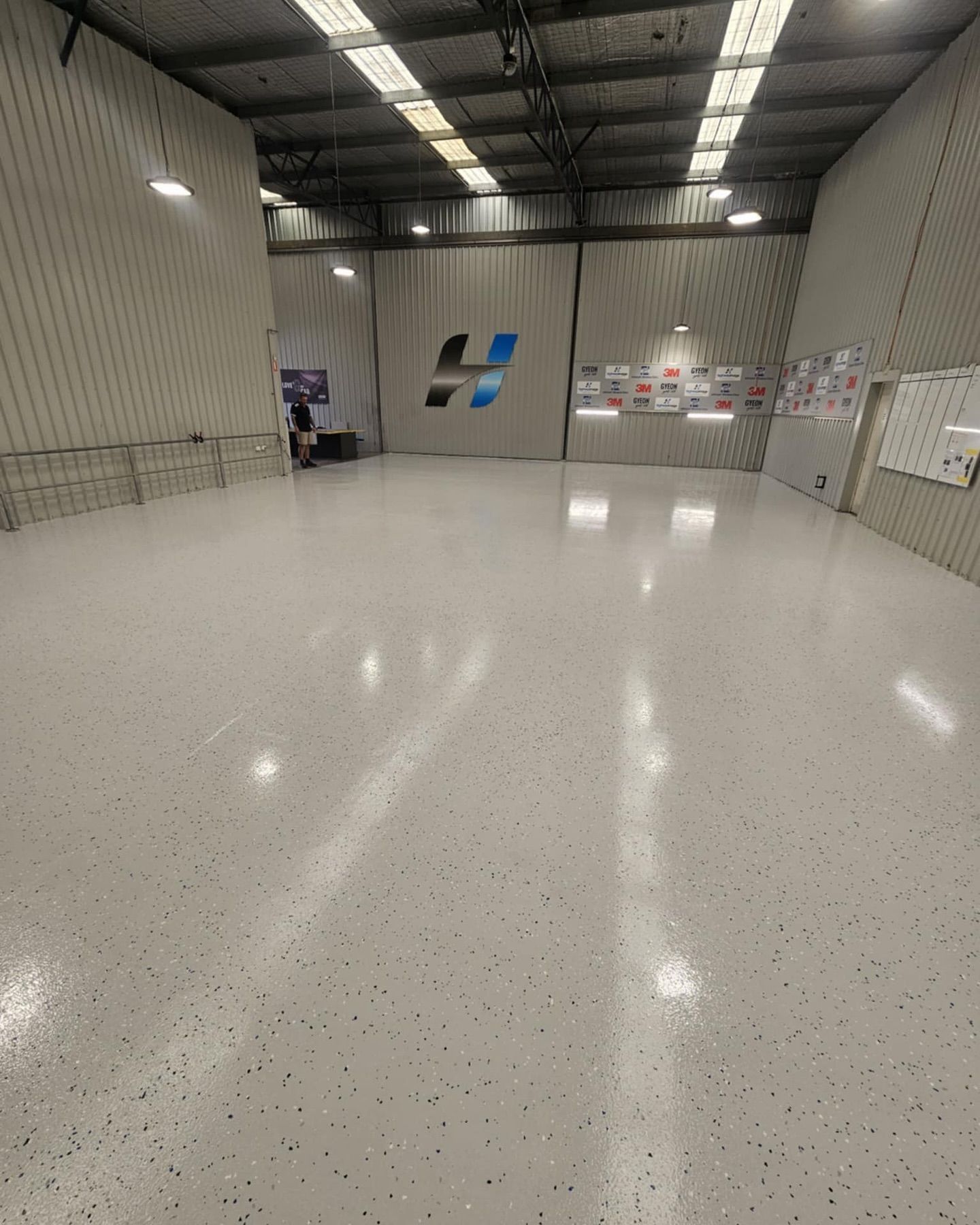 Empty warehouse with glossy white speckled floor, large logo on wall — Riverina Epoxy Flooring In Glenfield Park, NSW