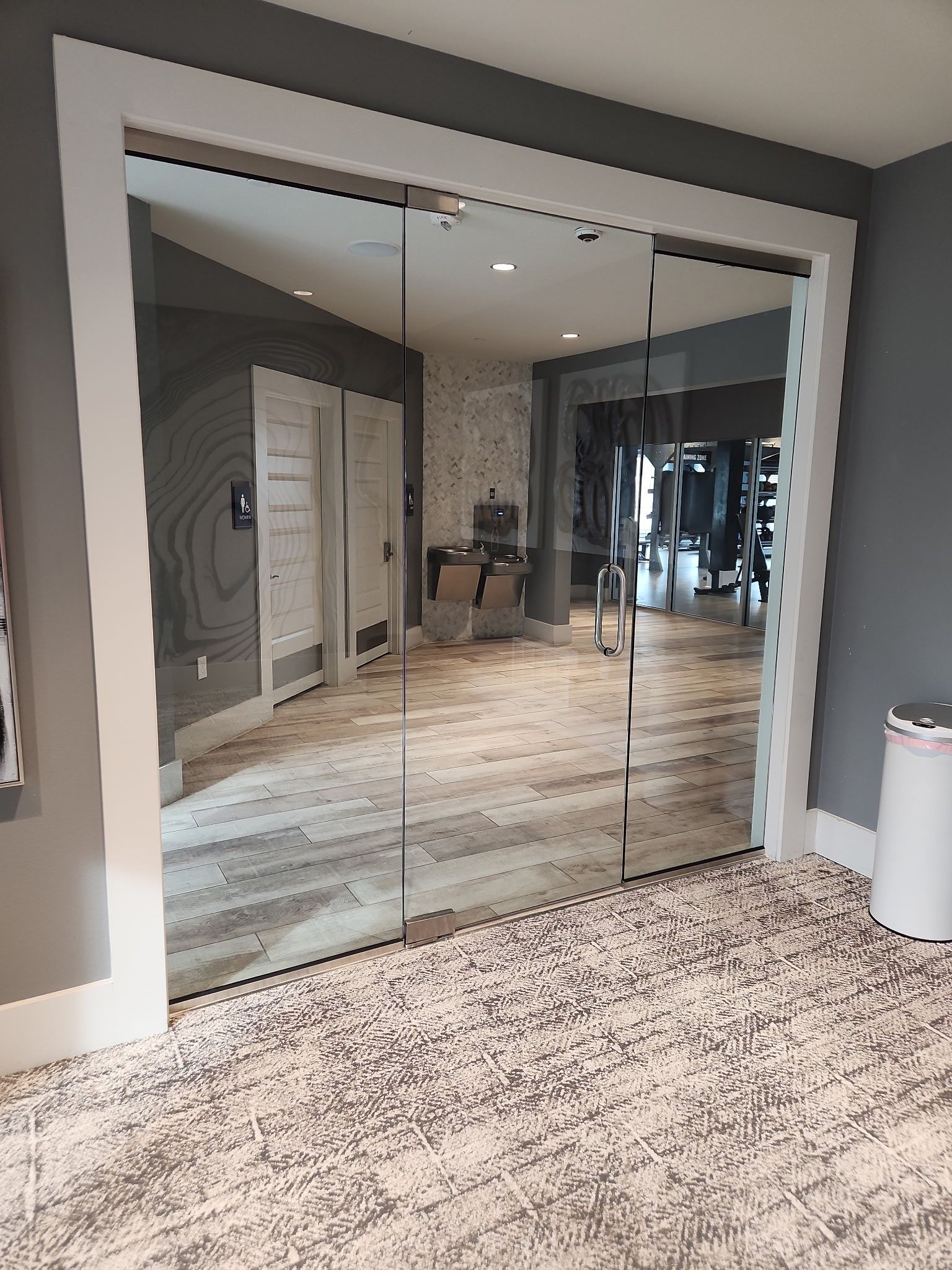 Glass lobby doors in a gray hallway, reflecting an interior hallway with carpet and lights.