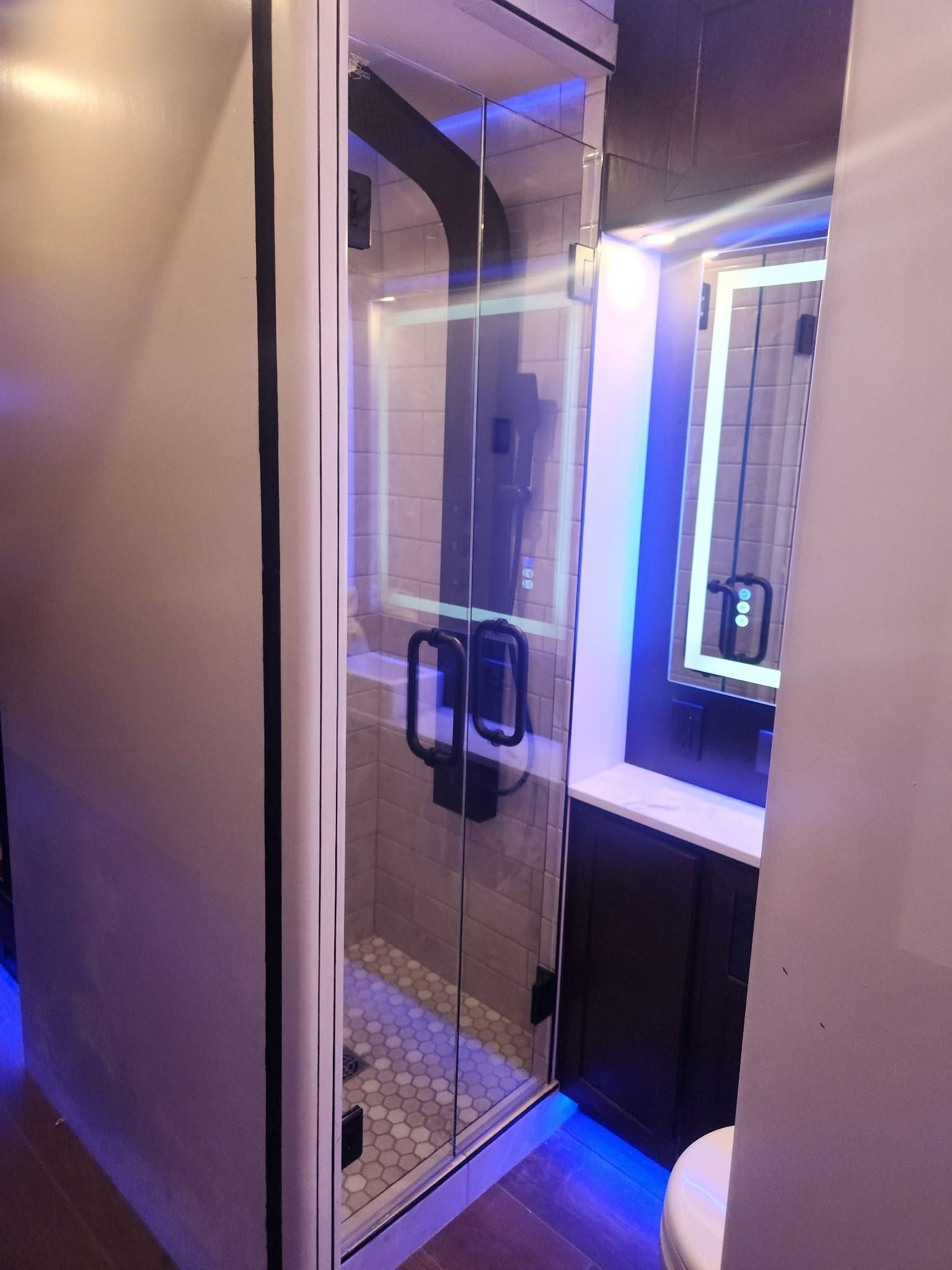 Modern bathroom shower with glass door, blue LED lighting, and vanity mirror visible through doorway