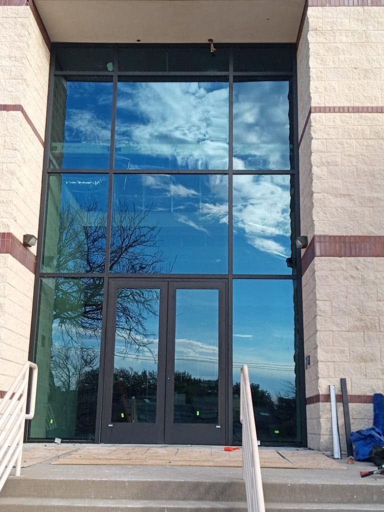 Glass entrance doors reflecting blue sky and clouds, framed by stone walls and handrails.