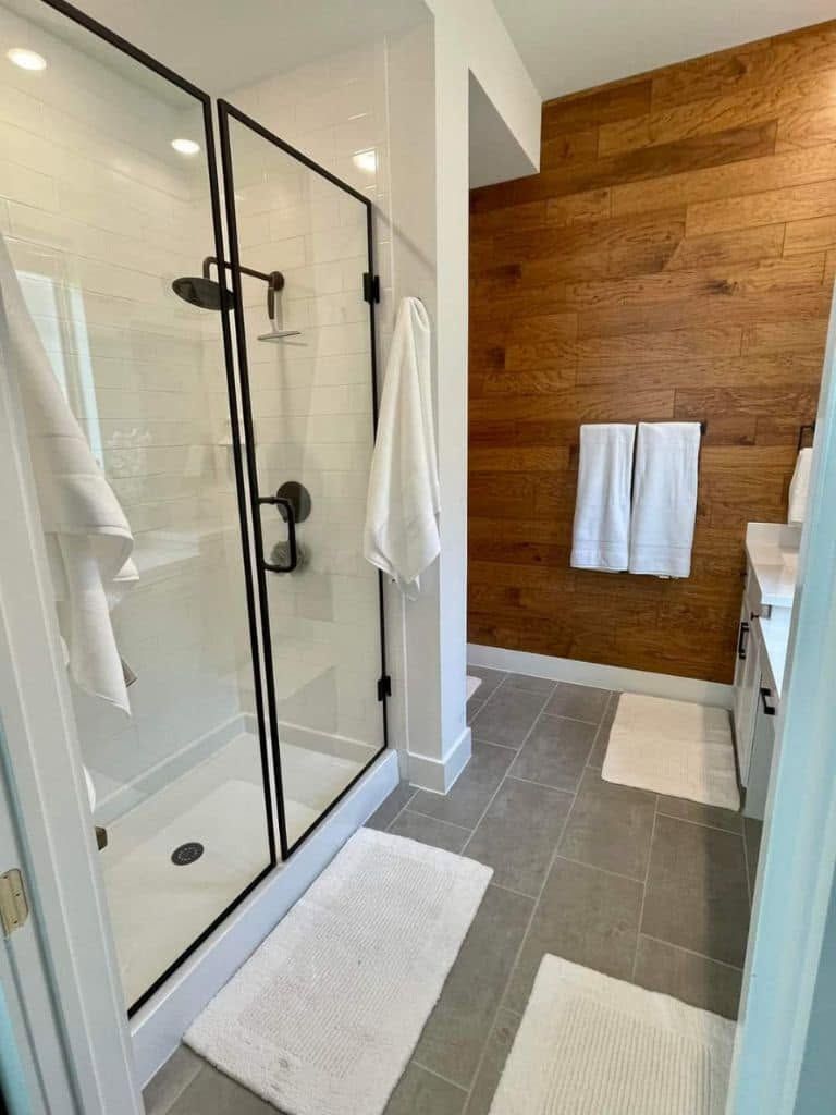 Modern bathroom with glass shower, white tub, gray tile floor, and wood accent wall with towels