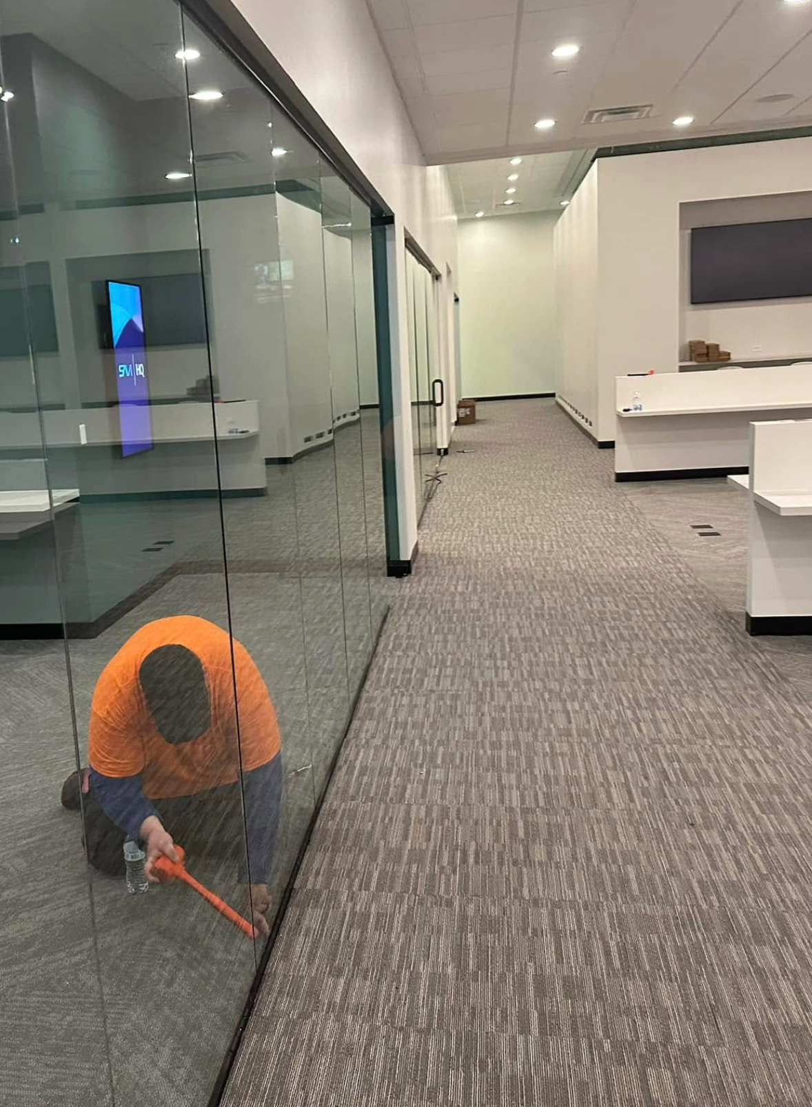 Worker vacuuming a carpeted office hallway beside glass meeting rooms
