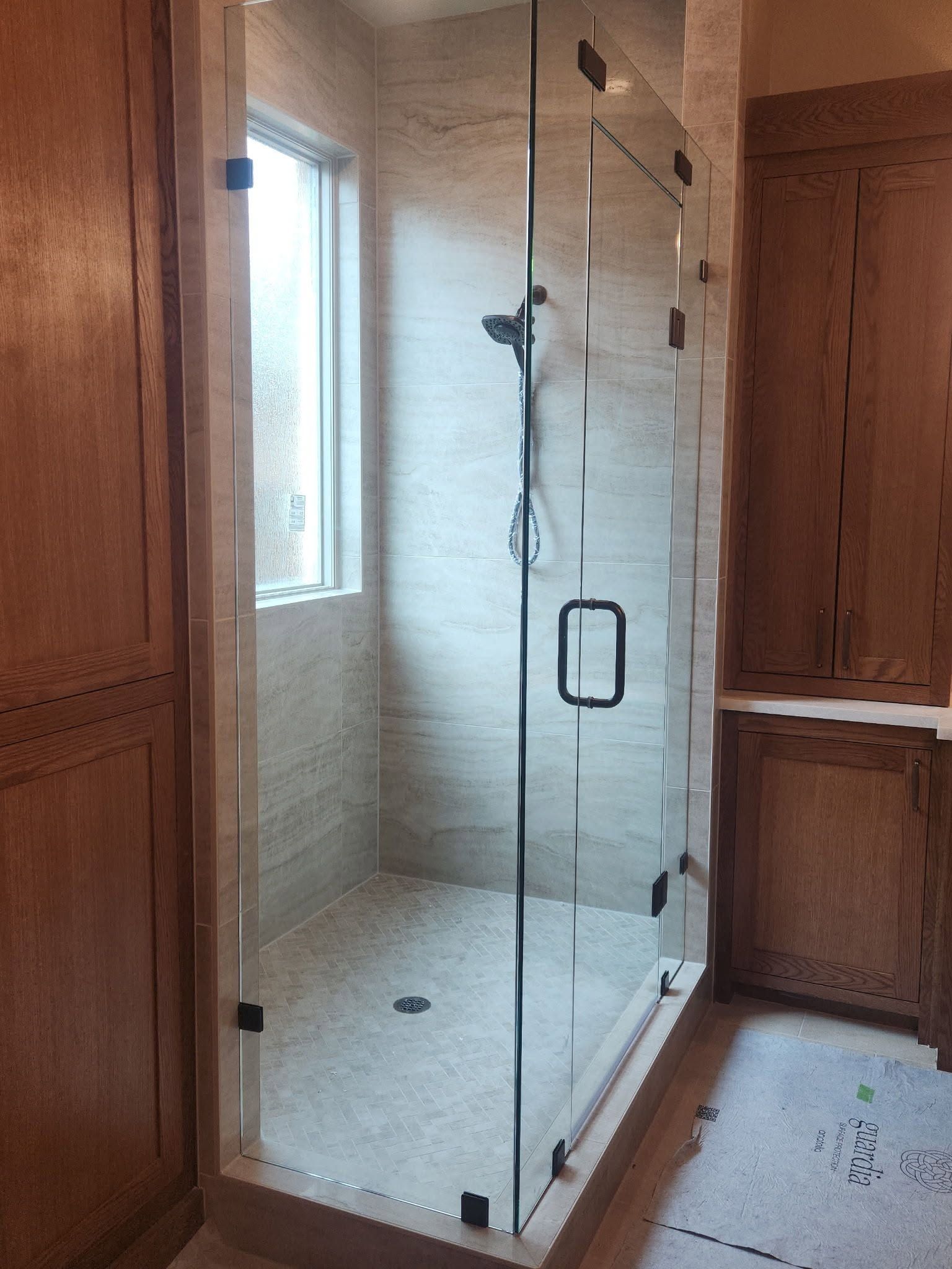 Glass shower enclosure in a wood-paneled bathroom with a window and tiled walls.