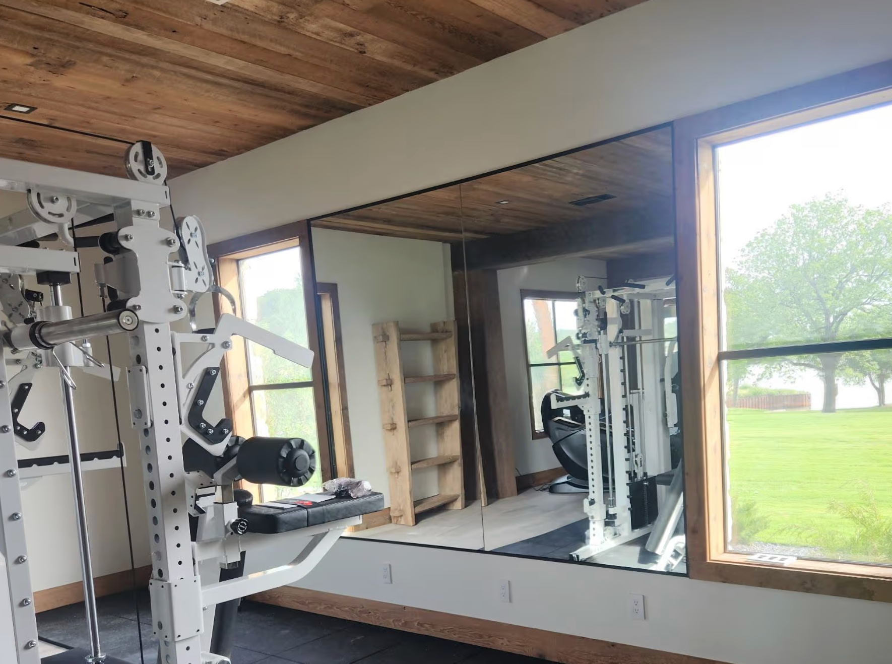 Home gym with cable machine, large wall mirror, and wood ceiling beside a bright window overlooking a lawn