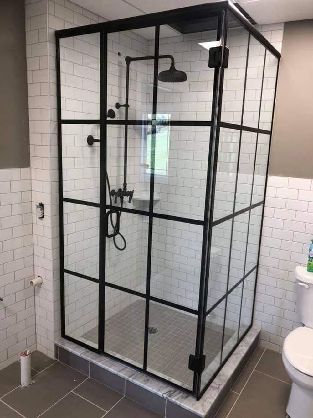 Black-framed glass shower enclosure in a tiled bathroom with shower fixtures and toilet nearby