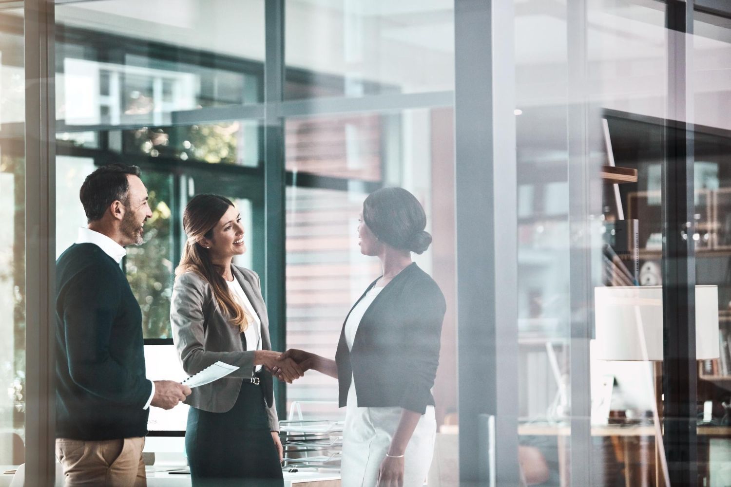 Three professionals in business attire talking in a modern office lobby, one holding papers and shaking hands.