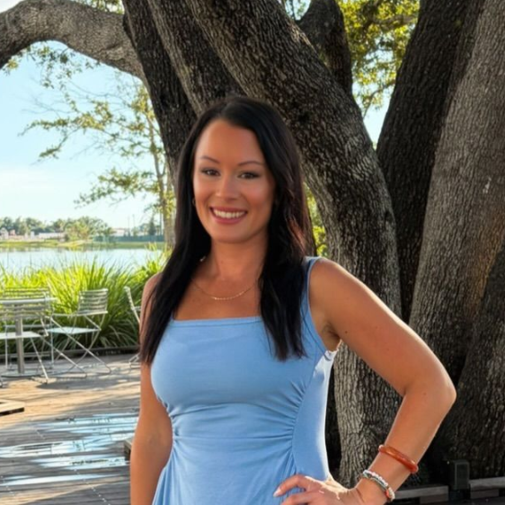 Woman in a light blue dress smiling outdoors beside a large tree by the water