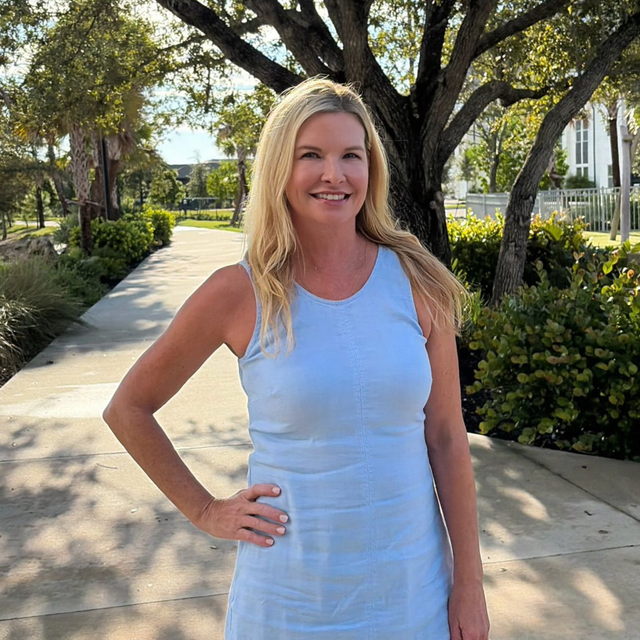 Woman in a light blue dress standing on a sunny sidewalk with trees and greenery behind her