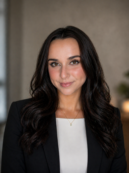 Professional headshot of a woman with long dark hair in a black blazer and white top against a neutral background