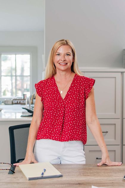 Woman in a red polka-dot blouse standing by a desk in a bright office, smiling.